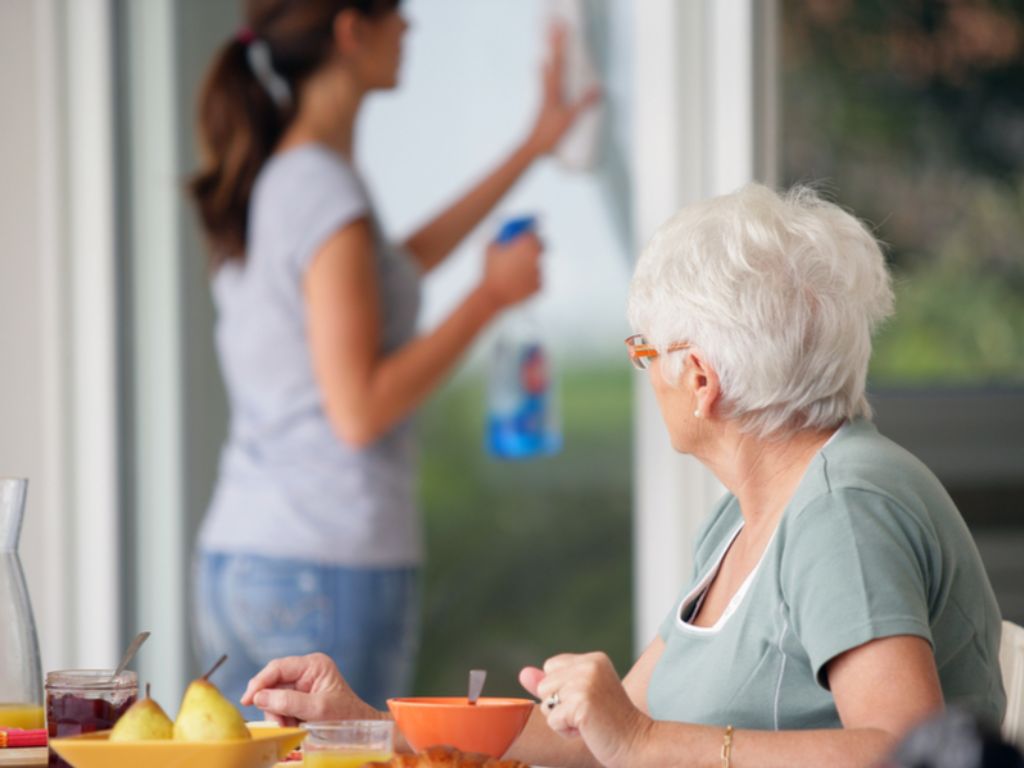 An elderly woman with white hair sitting outside at a table, looks back at cheerful care giver cleaning a sliding glass door. Light Housekeeping in Bowie