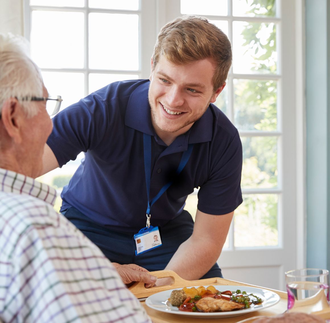 Light Housekeeping in Bowie A male caregiver serves a homemade meal to a senior man seated at the dining table, reflecting the thoughtful care and support that comes with services like light housekeeping in Bowie.