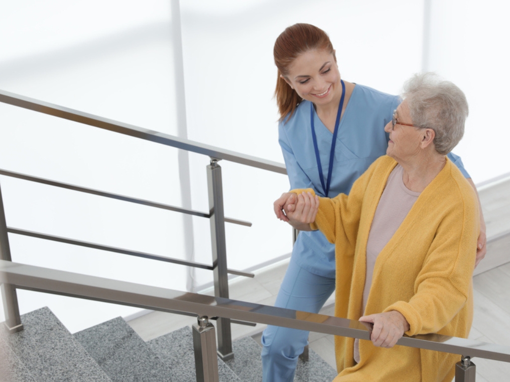 A cheerful caregiver holds the hand of an elderly woman assisting her as they walk up stairs. Personal Care in Bowie