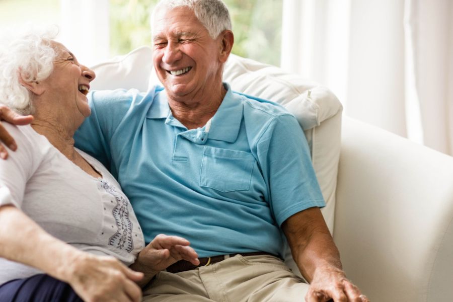 LTC An elderly couple sits closely on a couch, sharing a joyful moment and laughing together in a bright, cozy living room. The scene reflects the peace of mind and emotional well-being that can come from having reliable long-term care insurance.